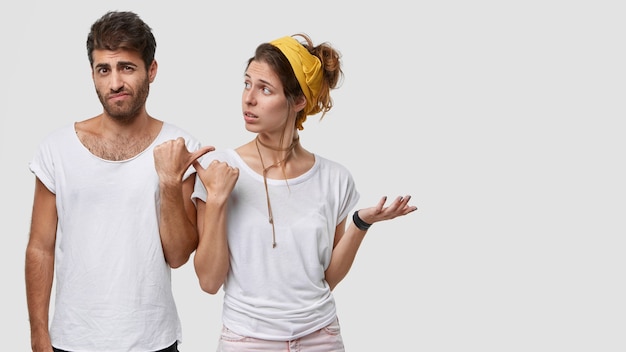 Free photo doubtful caucasian woman shruggs shoulders, looks at displeased man, wears white t shirt, model over  wall with free space on right side for your advertising content or promotional text