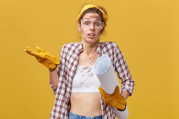 Doubtful female engineer doing manual work shrugging her shoulders with uncertainty. Dirty woman with protective eyewear, checkered shirt and yellow gloves holding blueprint.