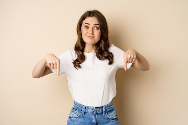 Doubtful girl pointing fingers down and smirking, looking uncomfortable while complaining, dislike smth, standing over beige background