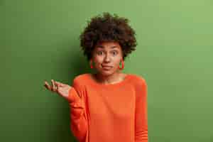 無料写真 疑わしい優柔不断な女性はためらいで手のひらを上げ、難しい質問または2つの選択肢に直面し、緑の壁に隔離されたオレンジ色のセーターとイヤリングを身に着けています。人、認識、態度