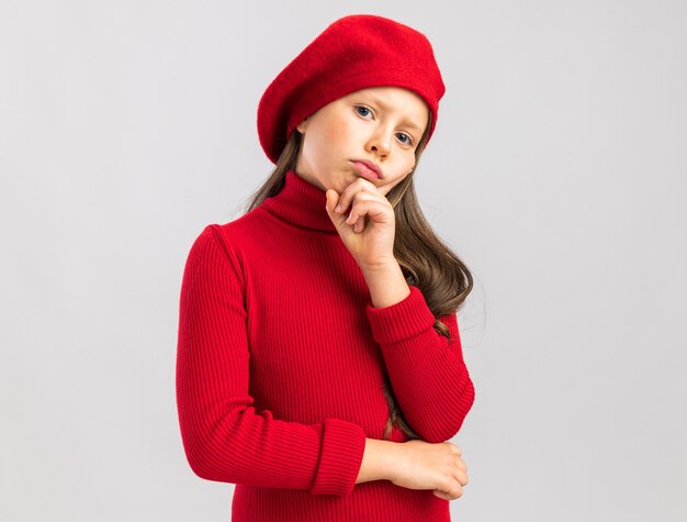 Doubtful little blonde girl wearing red beret keeping hand on chin  isolated on white wall with copy space