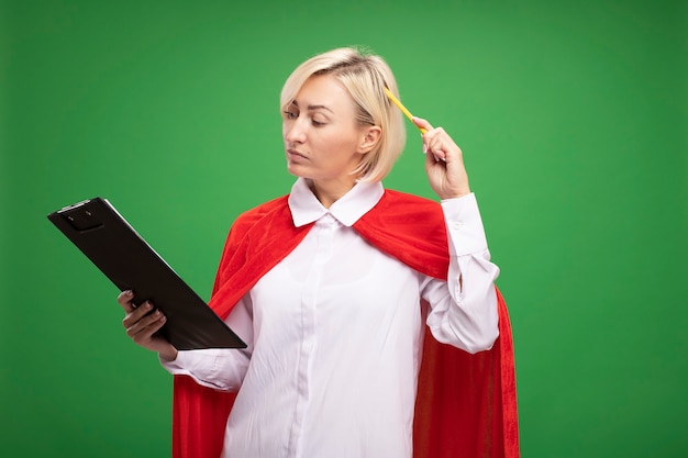 Doubtful middle-aged blonde superhero woman in red cape holding and looking at clipboard and touching head with pencil 