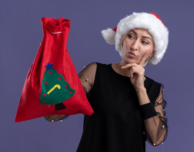 Doubtful middle-aged blonde woman wearing christmas hat holding christmas sack looking at it keeping hand on chin isolated on purple wall