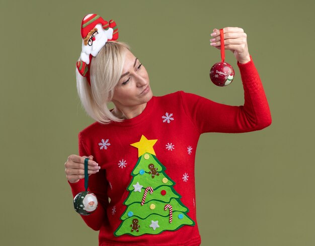 올리브 녹색 벽에 고립 된 하나를 찾고 크리스마스 싸구려를 들고 산타 클로스 머리띠와 크리스마스 스웨터를 입고 의심스러운 중년 금발의 여자