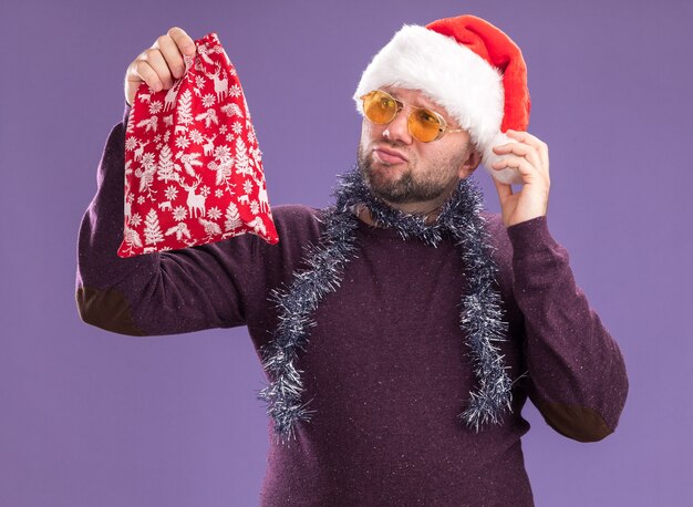 Doubtful middle-aged man wearing santa hat and tinsel garland around neck with glasses holding and looking at christmas gift sack grabbing hat isolated on purple background