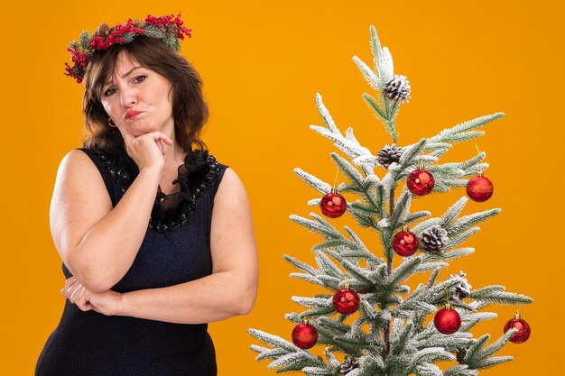 Doubtful middle-aged woman wearing christmas head wreath and tinsel garland around neck standing near decorated christmas tree