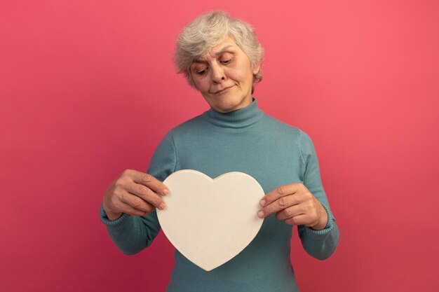 Free photo doubtful old woman wearing blue turtleneck sweater holding and looking at heart shape isolated on pink wall