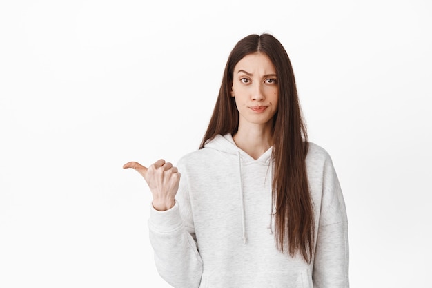 Doubtful and skeptical teenage girl frowning, looking with judging face and pointing aside at left logo banner, showing something bad or average, lame promo, white wall