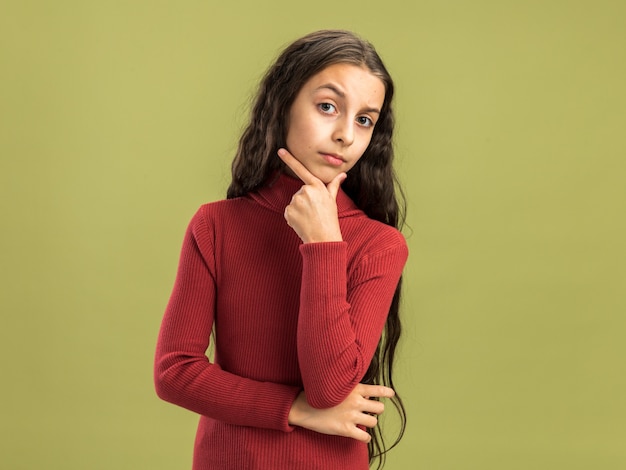 복사 공간이 있는 올리브 녹색 벽에 격리된 카메라를 보고 턱에 손을 대고 있는 의심스러운 10대 소녀