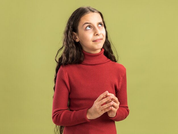 Doubtful teenage girl keeping hands together looking up isolated on olive green wall with copy space