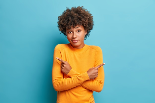 Doubtful woman crosses arms and points at different sides hesitates between two items or variants wears orange jumper poses against blue wall needs help with decision stands indoor