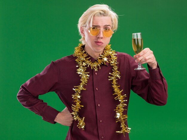 Doubtful young blonde man wearing glasses with tinsel garland around neck holding glass of champagne keeping hand on waist  isolated on green wall