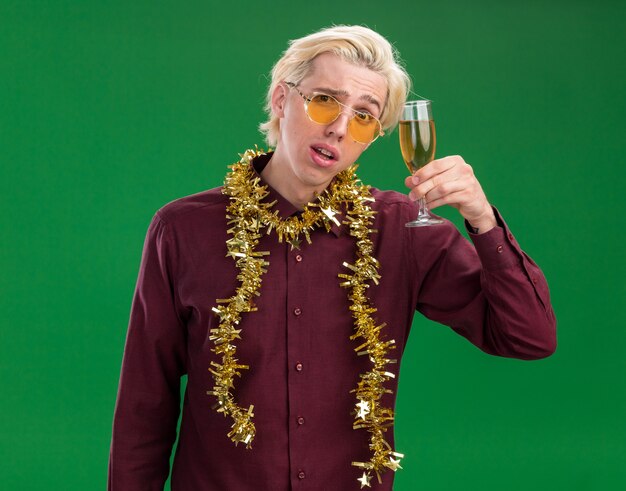 Doubtful young blonde man wearing glasses with tinsel garland around neck holding glass of champagne near head looking at camera isolated on green background