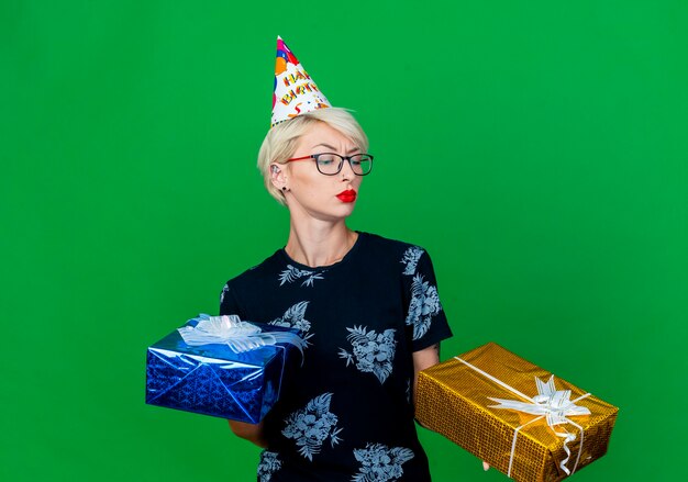 Doubtful young blonde party girl wearing glasses and birthday cap holding gift boxes looking at one of them isolated on green background