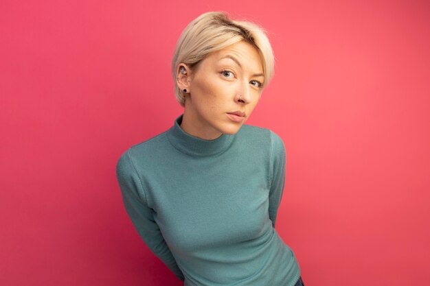 Doubtful young blonde woman looking at front keeping hands behind back isolated on pink wall with copy space
