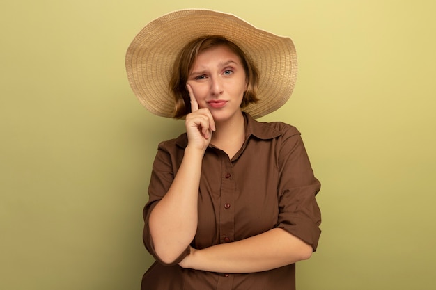 Dubbiosa giovane donna bionda che indossa cappello da spiaggia mettendo la mano sul viso guardando la parte anteriore isolata sulla parete verde oliva con spazio copia