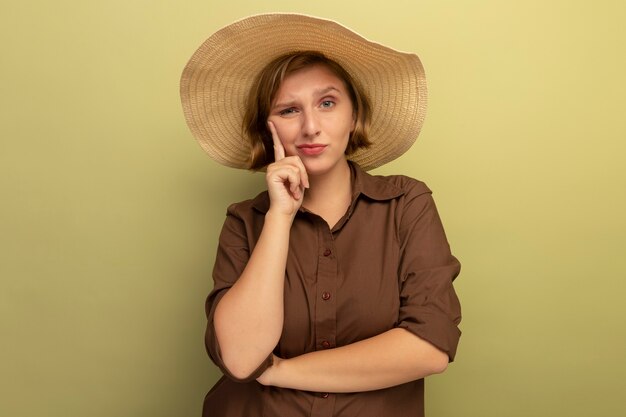 해변 모자를 쓴 의심스러운 젊은 금발 여성이 복사 공간이 있는 올리브 녹색 벽에 고립된 정면을 바라보는 얼굴에 손을 대고 있습니다.