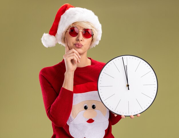올리브 녹색 배경에 고립 된 측면에서 찾고 턱에 손을 유지 시계를 들고 안경 크리스마스 모자와 산타 클로스 크리스마스 스웨터를 입고 의심스러운 젊은 금발의 여자