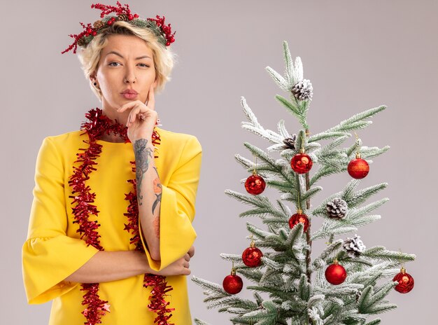 Doubtful young blonde woman wearing christmas head wreath and tinsel garland around neck standing near decorated christmas tree looking at camera keeping hand on chin isolated on white background