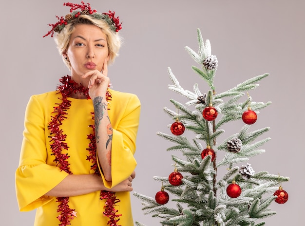 Doubtful young blonde woman wearing christmas head wreath and tinsel garland around neck standing near decorated christmas tree looking  keeping hand on chin isolated on white wall