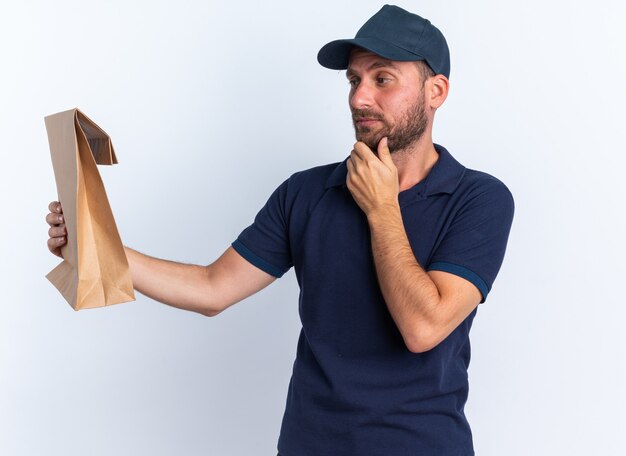 Foto gratuita dubbio giovane fattorino caucasico in uniforme blu e berretto che tiene la mano sul mento allungandosi e guardando il pacchetto di carta isolato sul muro bianco