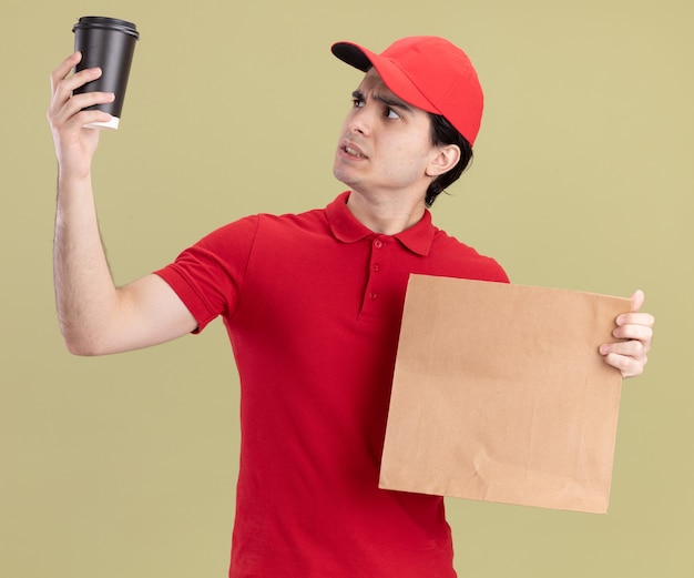 Doubtful young caucasian delivery man in red uniform and cap holding paper package and plastic coffee cup raising and looking at cup 