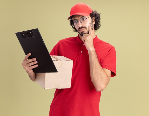 Doubtful young caucasian delivery man in red uniform and cap wearing glasses holding cardbox and clipboard looking at clipboard keeping hand on chin isolated on olive green wall