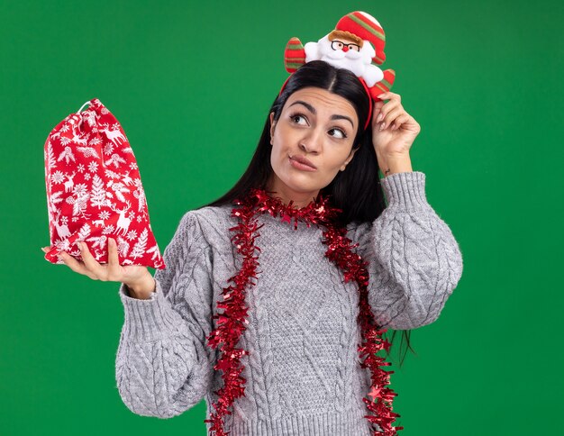 Doubtful young caucasian girl wearing santa claus headband and tinsel garland around neck holding christmas gift sack touching headband looking at side isolated on green background