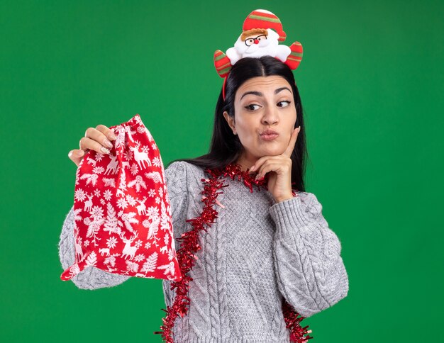 Doubtful young caucasian girl wearing santa claus headband and tinsel garland around neck holding and looking at christmas gift sack keeping hand on chin isolated on green background