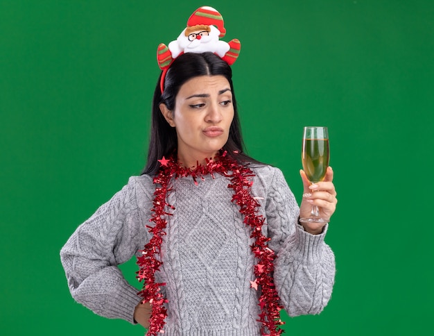 Doubtful young caucasian girl wearing santa claus headband and tinsel garland around neck keeping hand on waist holding and looking at glass of champagne isolated on green wall