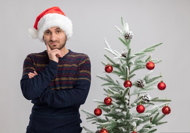 Doubtful young caucasian man wearing christmas hat standing near christmas tree  keeping hand on chin isolated on white wall