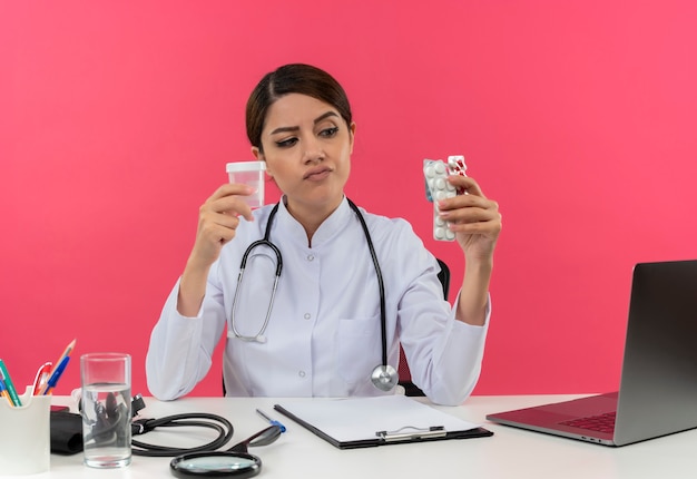 Сомнительная молодая женщина-врач в медицинском халате и стетоскопе, сидящая за столом с медицинскими инструментами и ноутбуком, держащая и смотрящая на медицинские препараты и мензурку, изолированную на розовой стене