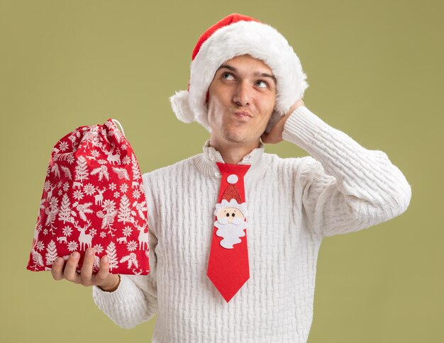 クリスマスの帽子とサンタクロースのネクタイを身に着けている疑わしい若いハンサムな男は、オリーブグリーンの壁に隔離された側を見て頭に手を置いてクリスマス袋を保持しています