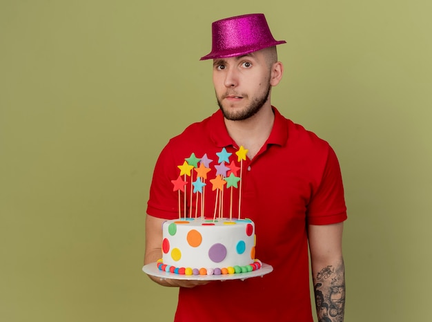 Doubtful young handsome slavic party guy wearing party hat holding birthday cake looking at camera isolated on olive green background with copy space