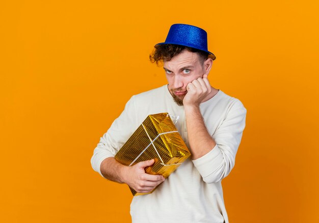 Doubtful young handsome slavic party guy wearing party hat holding gift box putting hand on face looking at camera isolated on orange background with copy space