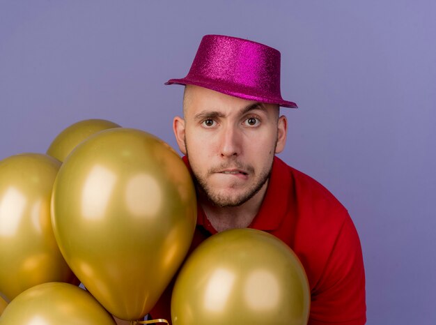 Doubtful young handsome slavic party guy wearing party hat standing behind balloons looking at camera biting lip isolated on purple background