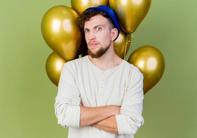 Doubtful young handsome slavic party guy wearing party hat standing with closed posture in front of balloons looking at front isolated on olive green wall