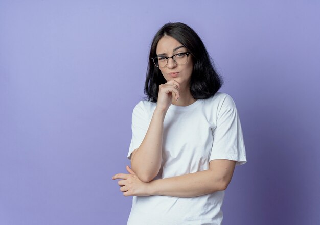 Doubtful young pretty caucasian girl wearing glasses standing with closed posture and keeping hand on chin isolated on purple background with copy space