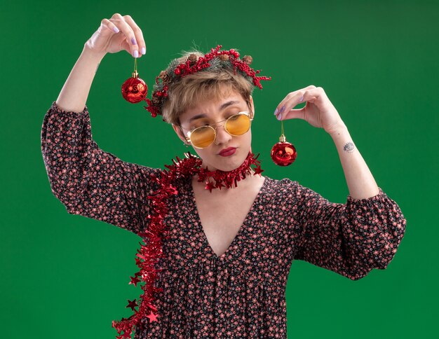 Foto gratuita dubbioso giovane bella ragazza che indossa la corona della testa di natale e la ghirlanda di orpelli intorno al collo con gli occhiali che tengono le palline di natale guardando uno di loro isolato su priorità bassa verde