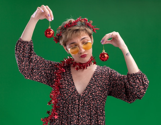 Сомнительная молодая красивая девушка в рождественском венке и гирлянде из мишуры на шее в очках с рождественскими шарами, глядя на одну из них, изолированную на зеленом фоне