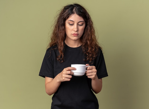 Dubbiosa giovane donna graziosa che tiene una tazza di tè guardandola dentro isolata sulla parete verde oliva con spazio per le copie