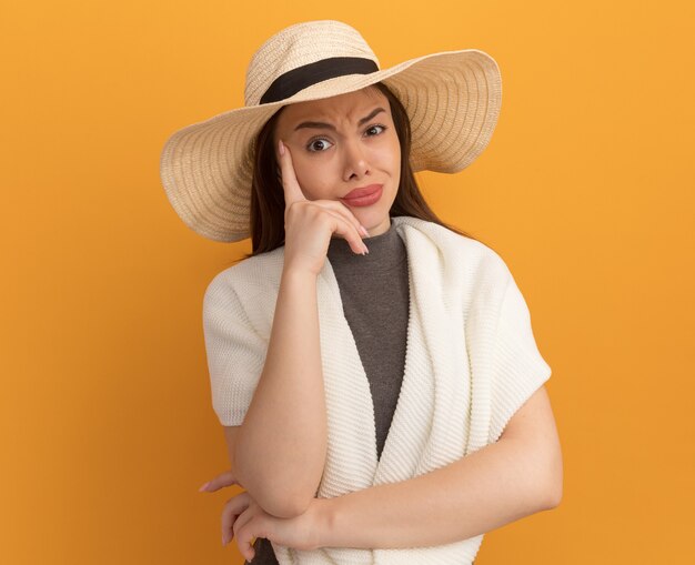 Doubtful young pretty woman wearing beach hat keeping hand on face 