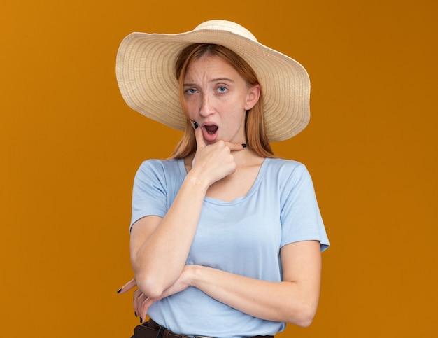 Foto gratuita dubbioso giovane ragazza rossa dello zenzero con le lentiggini che indossa il cappello da spiaggia tiene il mento e guarda la telecamera sull'arancio