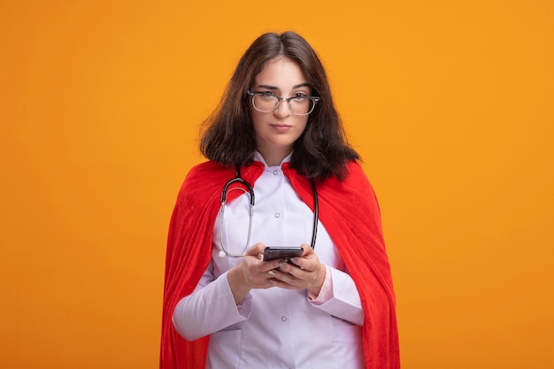 Doubtful young superhero woman in red cape wearing doctor uniform and stethoscope with glasses holding mobile phone looking at front isolated on orange wall with copy space