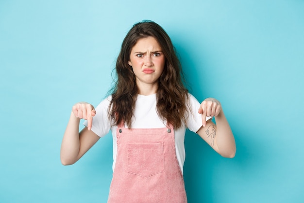 Doubtful young woman frowning and cringe from something bad, pointing fingers down at strange thing, having hesitations, blue background.