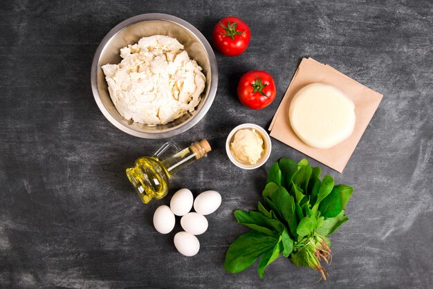 Dough, oil, cheese, tomatoes, eggs, greens over grey wooden surface