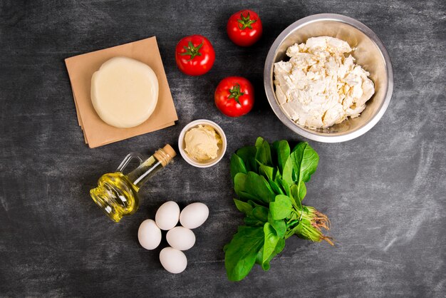 Dough, oil, cheese, tomatoes, eggs, greens over grey wooden surface