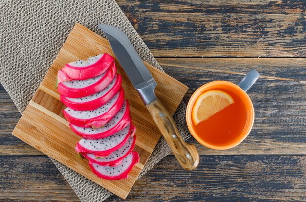 Dragon fruit with tea, cutting board, knife on wooden table, flat lay.