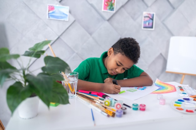 Drawing, pleasure. Happy cute dark-skinned school-age boy in green tshirt sitting at table enthusiastically drawing in room with easel and paintings on wall