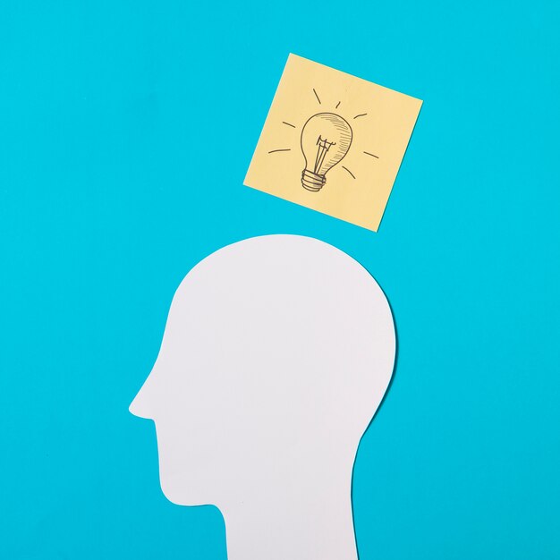 Free photo drawn light bulb icon on sticky note over the paper cut out head against blue background